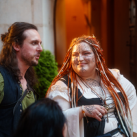 Two adults in costume converse outside a stone building; the woman has face paint and colorful dreadlocks, the man wears a green shirt with a vest.