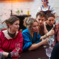 Group of friends seated at a long table in a rustic setting, chatting and holding wine glasses during a casual indoor gathering.