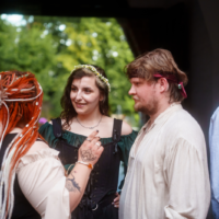 Group of four people in period-costume clothing chatting on a stage, with a woman wearing a flower crown at center smiling.