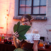 Woman in elf ears, off-shoulder white top and green skirt, holds a large leaf in front of her face at an outdoor bar.