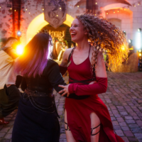 Two women dancing hand in hand at an outdoor party under warm string lights, one in a red slit dress, the other in black with purple-tinted hair.