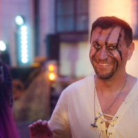 Smiling man with black face paint and goatee in a light beige pirate-style shirt at a nighttime outdoor festival.