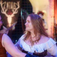 Two women in historical costumes dancing together at a medieval-style event; a stag banner is visible in the background.
