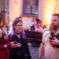 Three festivalgoers in medieval/fantasy costumes chat outdoors at dusk, warm lights glowing on a stone building behind them.