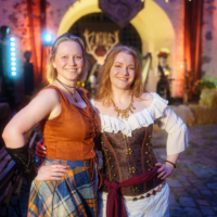 Two smiling women in medieval costumes posing together at a festival, with warm string lights and an arched background behind them.