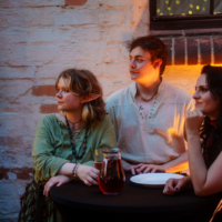 Three friends sit around a tall round table outside a brick wall, with a carafe of red drink in the center, bathed in warm orange light.
