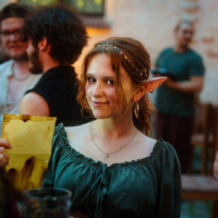 Young woman with elf ears at a social gathering, holding a yellow menu and smiling at the camera.