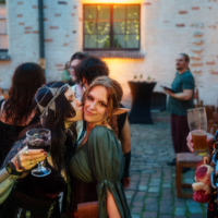 Two women in fantasy costumes share a kiss on the cheek at an outdoor, medieval-themed party, holding drinks as others mingle.