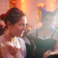 Woman in a lavender medieval gown holding a staff and wearing a delicate crown at a fantasy-themed party; a masked costumed guest is blurred in the background.