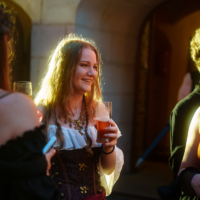 Woman in a corset and white blouse holding a glass of beer, smiling at an outdoor event.