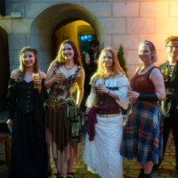 Five adults in Renaissance costumes posing with drinks outside a stone building, under warm lighting near an arched doorway.