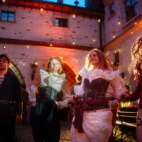 Group of four in Renaissance costumes dancing in a cobblestone courtyard under warm string lights at night.