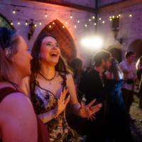 Young woman in a corseted dress dancing and laughing with a friend at a lantern-lit courtyard party.