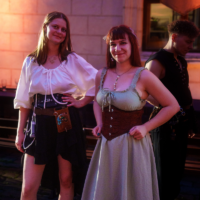 Two women posing in period-costume outfits under warm ambient lights, one in a white blouse and black skirt with a belt and pouches, the other in a light blue dress with a brown corset and necklace.